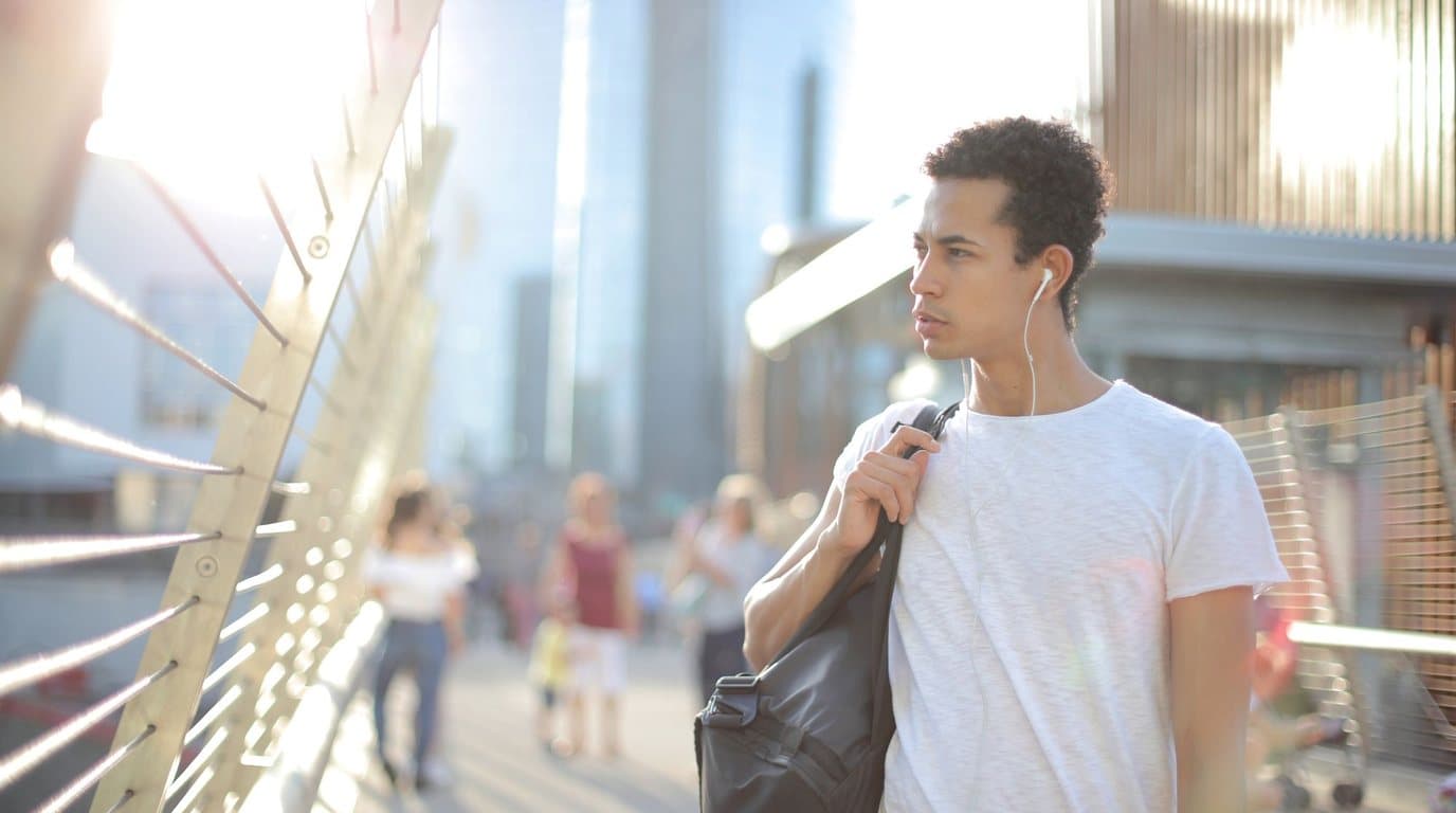 A young man walking through a sunny city street wearing earbuds